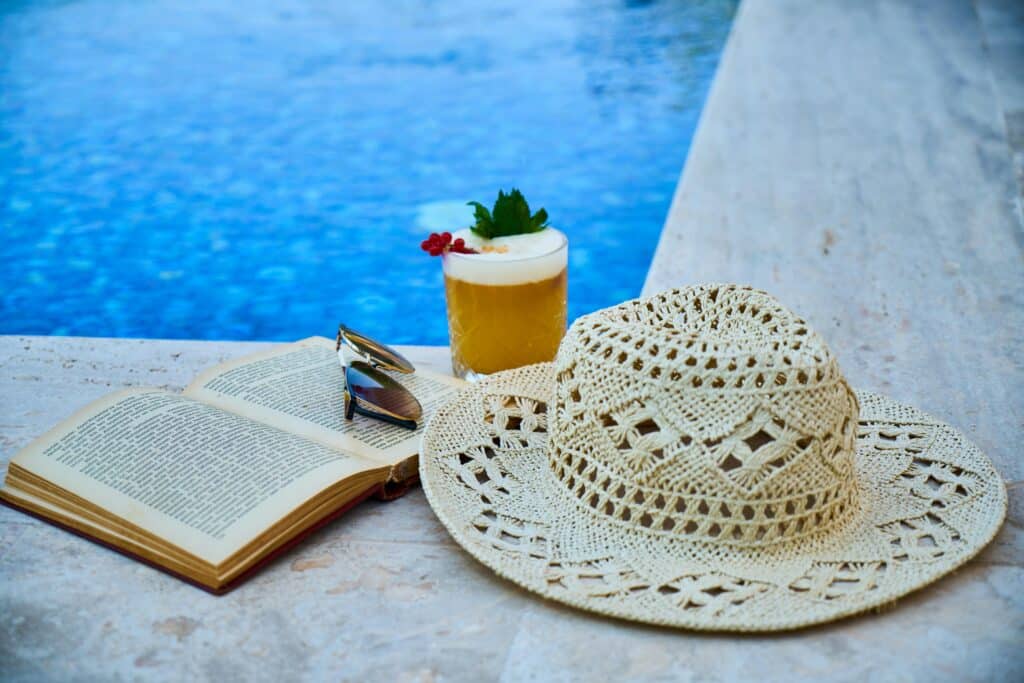 pexels-enginakyurt-lunette de soleil et chapeau et livre et cocktail au bord d'une piscine-8 vêtements indispensables pour slay en vacances